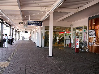 外の白い鉄骨の屋根の下。右手に待合室の出口。左手は駅前の回廊で地面はくすんだとび色のタイル地になっている。
