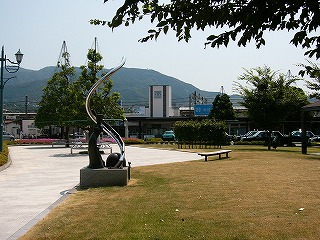 右手に芝生、左手には石畳の歩く道。向こうには三角の木2本と噴水、花のある花壇。