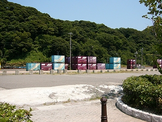 アスファルトの敷地の向こうにコンテナ積み。すぐ後ろに横長のすごく低い山。