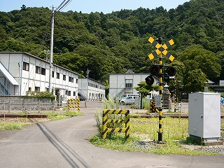後ろに低い山、左に白いプレハブで2階建ての建物。手前に踏み切り。