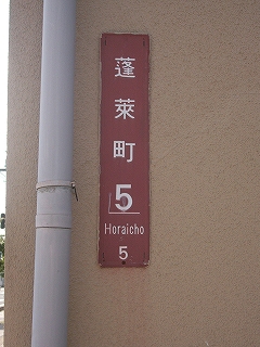 縦長の看板でエンジ色の背景に白地で「蓬莱町5」と書かれている。