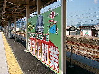 背景は黄緑で、丸っぽい白い字に赤の影をつけた「直流化開業」の文字が書かれいくつかのイラストも描かれた宣伝の看板。高さ170cmぐらいで横に長い。