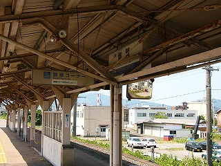 上屋の下。鉄骨に吊り下げられた駅名標のすぐ前に暗いベージュの古びた鉄骨が走り、読みにくくなっている。左向こうには風除け2つ。右奥には駐車場が見える。