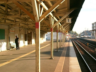 ホームの縁からホームと線路の伸び行くのを見て。ホームでは、ひび割れた白い木の柱が並び上屋を支えている。