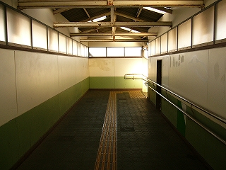 跨線橋内部。両側の壁の下の部分は緑色に塗られ、それより上の部分は白色。それより上の部分と屋根の間には不透明な明り取りのアクリル板のようなものが取り付けられた窓框が並んでいて、弱い光が入ってきている。床面は濃い緑色の正方形のパネルが敷き詰められている。