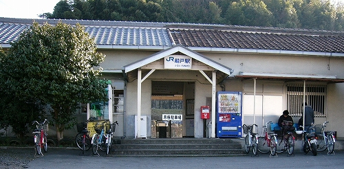 出入口に三角屋根の付いた白い駅舎。駅舎前に何台かの自転車、そして木々。
