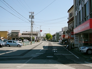 二車線の道路、個人的なビル、民家。自動車は一台も通っていない。