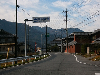 中央線のない道路。左に家一件、右に家々。上に加太駅への案内看板が出ている。