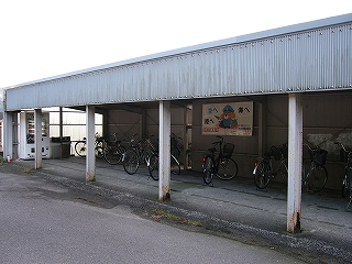 灰色のトタン屋根を白い鉄骨で支えて作られた無骨な駐輪所。軒端には地面と垂直に灰色のトタンが40センチぐらいの長さで取り付けられている。