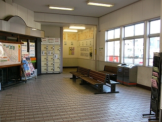 2色のレンガタイルを1段ごとに色を違えてはめ込まれた床の待合室。右手には外に面した窓が並び、タイル床の上には焦げ茶の長椅子が置かれている。待合室のスペースの左側は左斜めにカットされ、そこには閉店になった暗いガラス戸の入口がある。