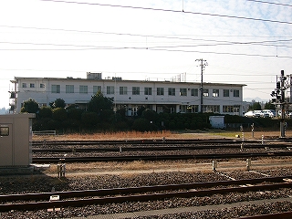 幅広い線路内の向こうに見える白い壁の二階建ての横にずいぶん長い無機質な建物。建物の前には緑が茂っている。