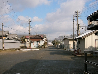 向こうから来て道が少し広く膨らんだ所にある駅前スペース。左手には倉庫やガレージ、民家などが見られる。回りには商店などはなく、古い住宅地の中、という雰囲気。