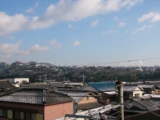 数々の民家の瓦屋根の向こうに斜面が樹木で覆われた丘陵地を見て。丘陵地上にも家々が立ち並んでいる。