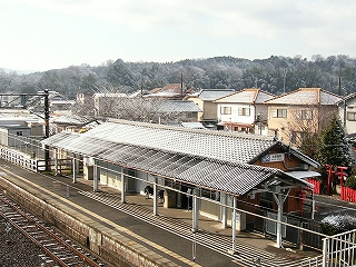 妻面から見て縦長である駅舎を斜めに見て。山型の上屋は駅舎の山型の瓦屋根と接している。