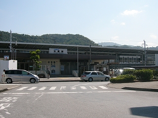 少し離れて駅舎を見て。目の前にロータリーの一部の太い道があり、それが右にカーブしながら駅舎前に続いている。