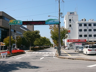 グリーンベルトのある道路の右車線を正面から見て。門形になった上部に一行の横長の電光掲示板が設置されていて、米原市のサイトのアドレスを流している。