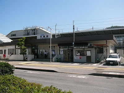 四方に張り出した焦げ茶の陸屋根とクリーム色の壁の平屋の駅舎。駅舎のすぐ前には幅の広めの歩道。そして、ロータリーの一部をなす道路が通っている。商店などはまったくない。