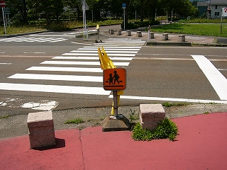 横断歩道の前に建てられた旗立て。黄色地に子供と大人が渡っている黒い図案の、標識のようなものが箱状になっていて、黄色い旗が入れられている。