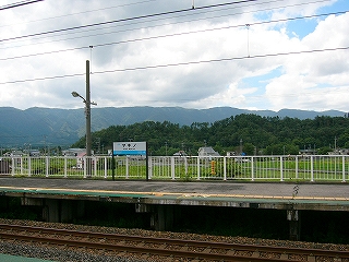 向かいのホームの上屋のない部分。背後に白い柵があり、その向こうに田と林、山が見える。