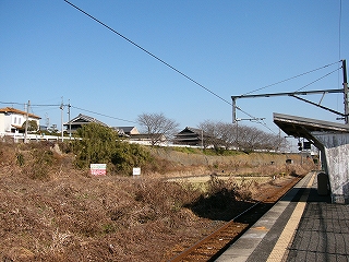 線路脇には山のような枯れ草の山と田んぼ。すぐ向こうには少し高い位置に住宅が並ぶ。
