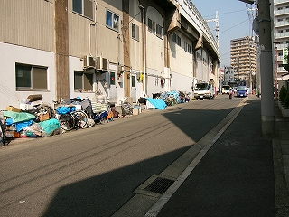 少し上り坂。左手に半球の効果があり、その高架下は壁が作られて倉庫になっている。それに沿って、道路上には種々雑多なものが寄せ集められている。青いビニールシートも目立つ。