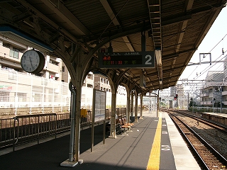 ホームの屋根の下。左側は日陰で右側は日が照っている。屋根から丸くて厚い時計と行き先案内が吊られ、ホームの上には二本足で立っている時刻表と背中合わせの3連の椅子。