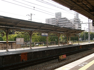 V字型の上屋を持つ下り線ホームを左斜めに見て。ホームの向こうの敷地には緑の茂った並木があり、その向こうにある静かなロータリーが見えにくくなっている。右手奥にはマンションが建っている。