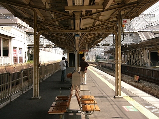 ホームの上屋下の真中から眺めて。写真すぐ手前真中に薄茶色の3連椅子、その奥にゴミ箱、そしてクリーム色の柱。ずっと奥に跨線橋への階段が立ちはだかっているため、それより先のホームのようすは見えていない。