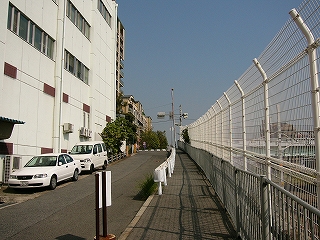 坂道の先に信号のある交差点がある。右手には低い位置線路があり、背の高い白い網の柵で遮られている。