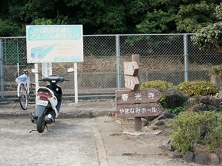 古いコンクリート面の上にとめられた白い自転車と白いスクーター。右に庭の低い植え込み。