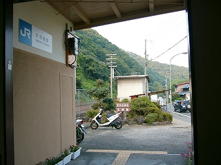 暗い駅舎の中から、扉のない大きな長方形の出口を通して、植え込みのような小さな庭とその前にあるバイクを正面に見て、右手に道路の端を見る。背景は線路を挟んだすぐ向こうにある緑の樹木の茂った山肌。