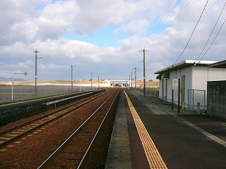 上屋のない長いホームに立って。左手に線路内、右斜めにホーム脇の信号機室がある。信号機室は白いコンクリートの平屋の建物。