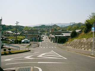 まっすぐ伸びる二車線の道。少し向こうでＴ字路にぶつかっている。沿道には建物が並ぶ。遠くに山並み。