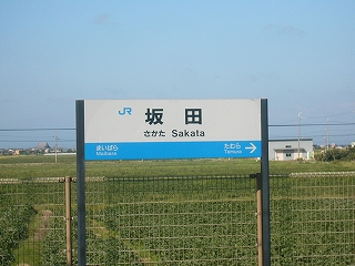 屋根のないホームに、白い網の柵に寄り沿って立つ駅名標。柵の向こうに緑の畑が広がる。