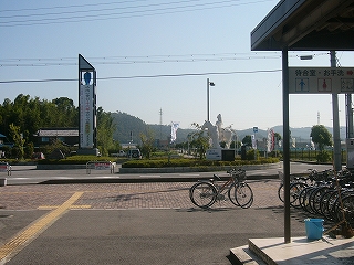 縁石とつつじの植え込みで造られた駅前の新しいロータリー。ロータリー内の左の方には植え込みの中に立っている塔が見える。撮影地点のすぐ右手には並んだ自転車と待合室の玄関の端が写っている。