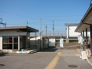 左にプレハブのような簡易な平屋の待合室、その奥に地下道の入口、その右となりに大きな時刻表がある。奥には駅のホームが高い位置に見え、右手にはコミュニティハウスの端が写っている。