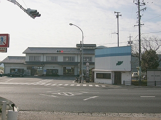 交差点に出て。奥には駅舎。道路を渡った右手に駅前にはふさわしくないような二階建ての波板の壁の小さな建物。二階部分に窓がない。