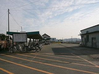 左手にほとんど平らに近い薄いＶ字型になった厚みのある屋根を持つ駐輪所を縦に見て。右手には広いアスファルトの敷地が広がる。その敷地の右端には線路が通っている。