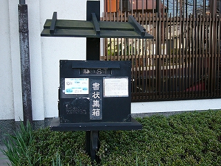 ほぼ立方体の黒色の箱の郵便ポスト。箱の後ろから棒が上に延びて、その棒に箱のための庇が付けられている。