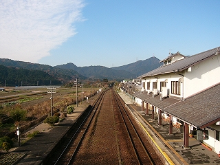 右手に白い壁で瓦葺の、二階建ての駅舎。真中にこげ茶色の線路内、左手には田畑が広がる。