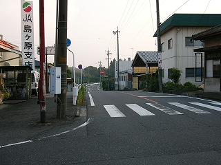 下りながら延びて向こうの先で旧カーブする道。すぐ左にはJA島ヶ原立て看板と長いポールに支えられた時計と建物、右手は簡易な工場。