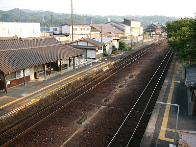 真中に焦茶色のバラストの目立つ構内。旧3線式で、中線が取り払われている。左に回廊つきの駅舎と下りホーム、右には上りホームとホーム上の緑の木