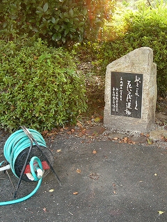 つつじの植え込みと水色の巻き取りホースと膝丈ぐらいのささやかな碑。