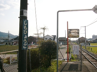 縦型の駅名標とミラー、田んぼと集落、夕日。