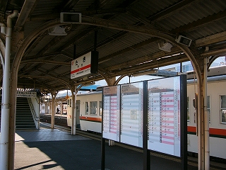 低めの上屋の下に立つ三つの時刻表と、その上付近に上屋から吊られた駅名標。