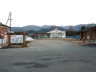 駐車場スペースとログハウス風のトイレ、丸太で組んだ案内板。遠くに取り囲むような山々。