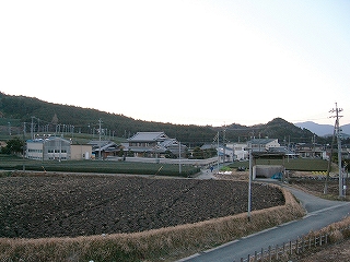 濃いこげ茶の土色の田んぼといくつかの田舎らしい大きな家と遠くに茶畑、山。
