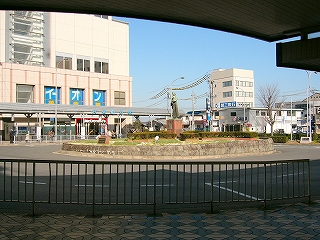 手前に柵、その向こうにロータリー。ロータリーの真中には植え込みがあり、銅像が立てられている。