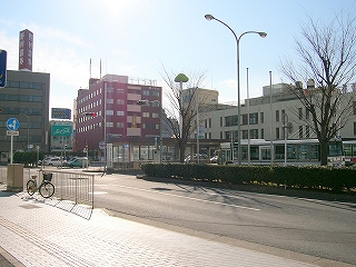 左斜めに二車線道路を見て。右隣にバスロータリー、そして駅舎。奥には6階建てのビルが２つ並んでいる。