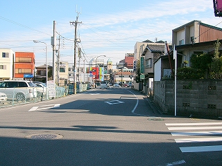 中央線のない、車の通りの少ない道路。左側に駐車場、右手に人家の並び。遠くに商業施設のようなものが見えている。